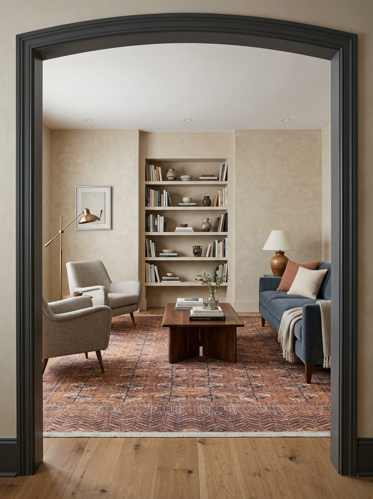 A cozy living room with the Earthy Enigma - Washable Rug - JR1937, ideal for high-traffic areas, two beige armchairs, a dark blue sofa, wooden coffee table, built-in shelves with books and decor, floor lamp, and light neutral walls seen through an arched doorway.