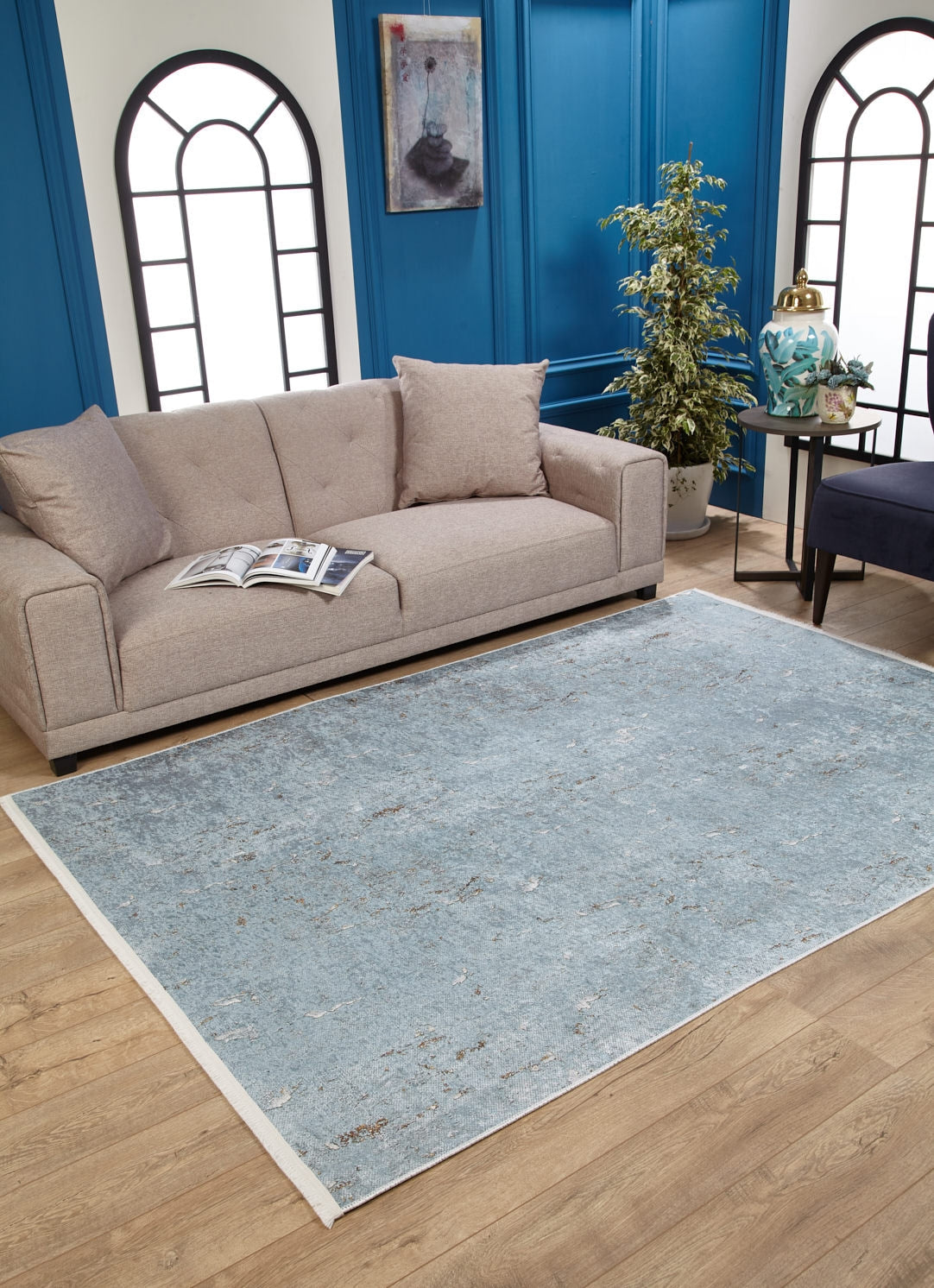 A modern living room with a beige sofa, open magazine, potted plant, blue paneled walls with arched windows and a painting. The light wood floor features the Earthy Sands - Washable Rug - JR1819 (Custom Size), ideal for high-traffic areas.