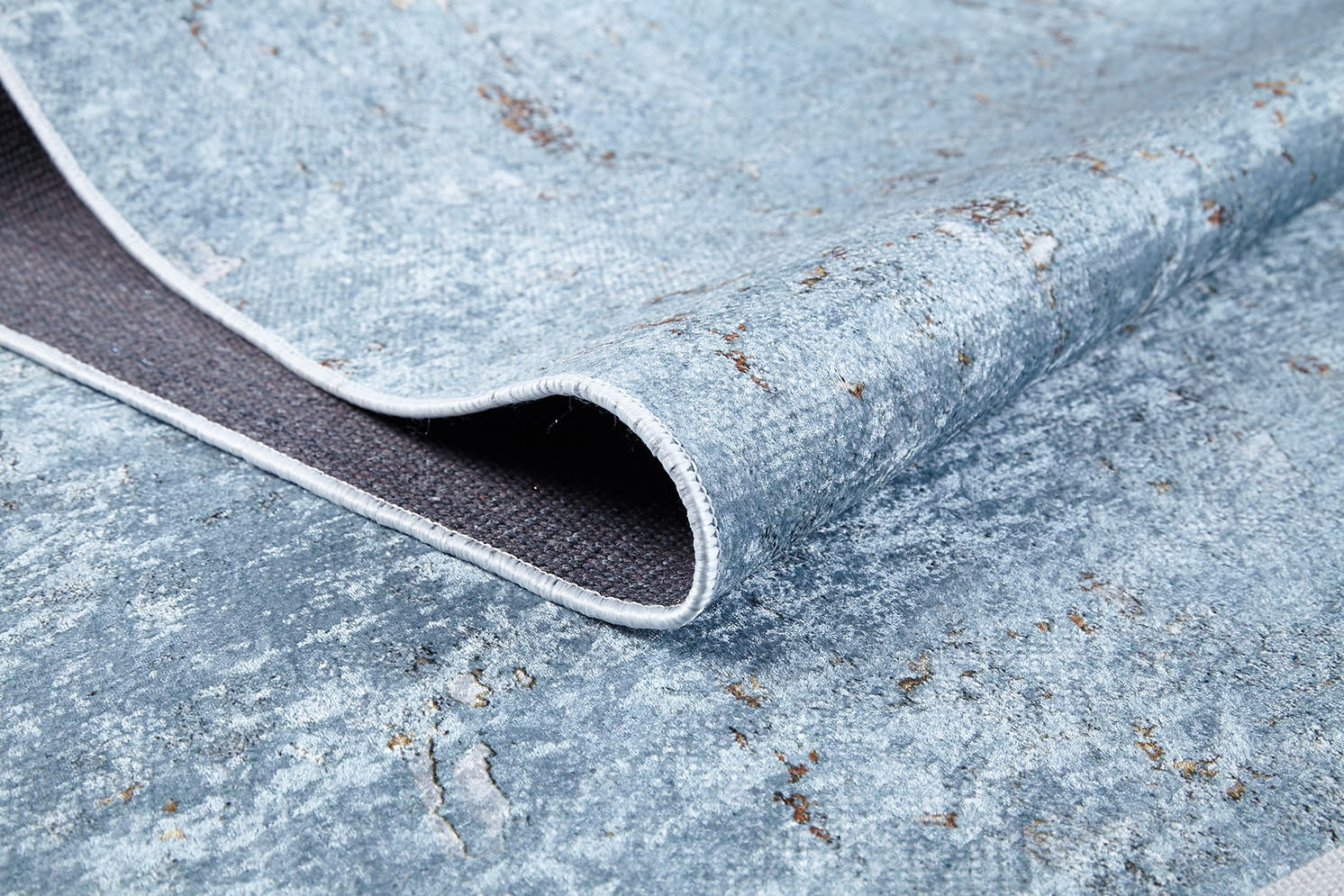 A close-up of the Earthy Sands - Washable Rug - JR1819 (Custom Size) shows its textured blue surface, brownish weathered accents, curled edge, and dark woven underside—perfect for high-traffic areas.