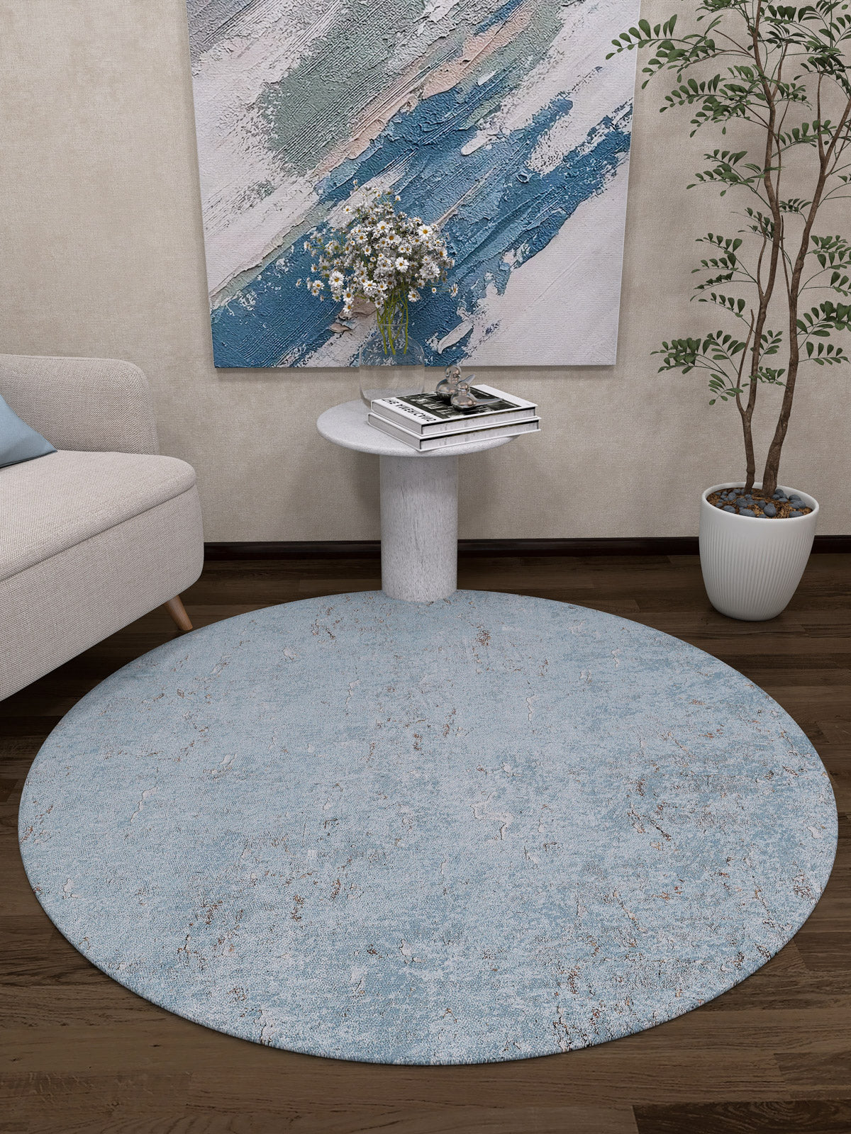 A modern living room features the Earthy Sands - Washable Rug - JR1819 (Custom Size), perfect for high-traffic areas, along with a small table with flowers and books, a white sofa, a potted plant, and an abstract blue and white wall painting.