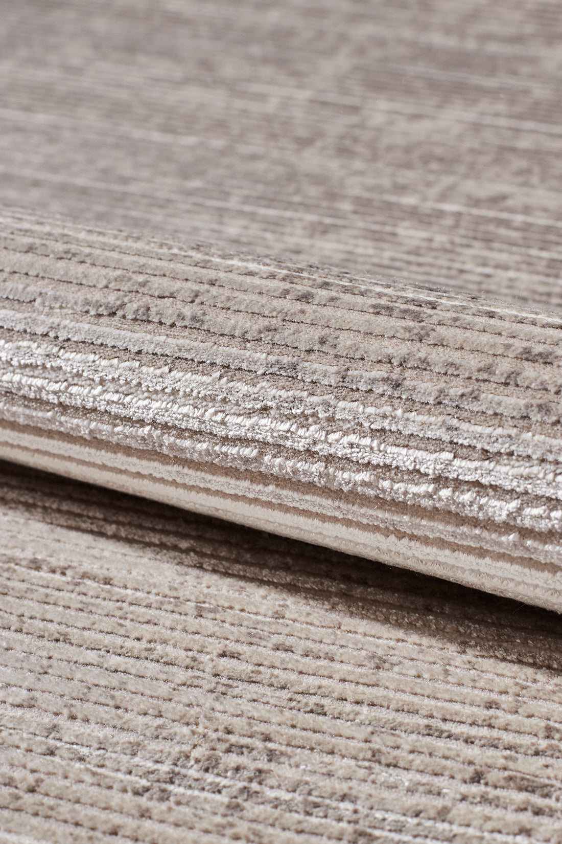Close-up of the Earthy Textures Neutral Rug - M497J in beige and light brown, featuring subtle horizontal lines and a soft, slightly shiny flat pile. The rug is partially folded, showing its thickness and detailed fabric texture.