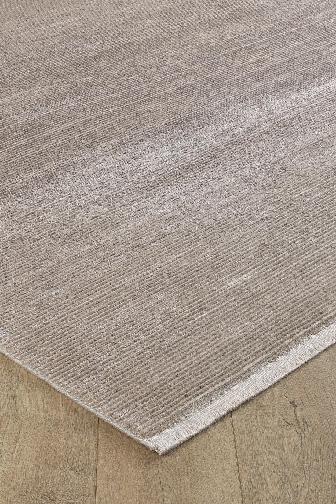 A close-up of the Earthy Textures Neutral Rug - M497J shows its beige flat pile with subtle horizontal lines and a slight sheen, placed on a light wood floor. One minimalist edge is finished with a short fringe.