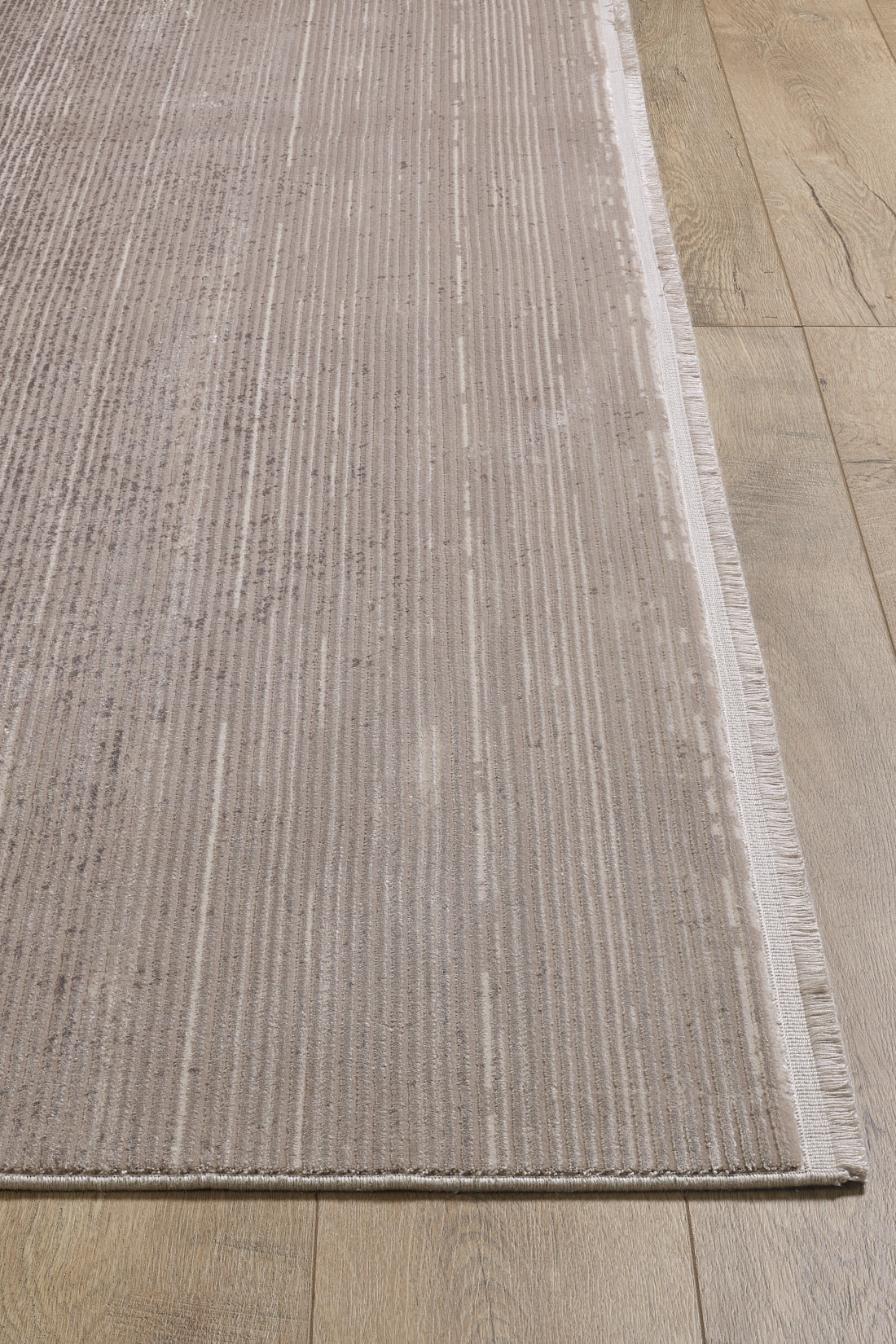 A close-up of the Earthy Textures Neutral Rug - M497J, a beige flat pile rug with subtle stripes, fringed edges on one side, and a modern machine-made design on light wood flooring—perfect for minimalist spaces.
