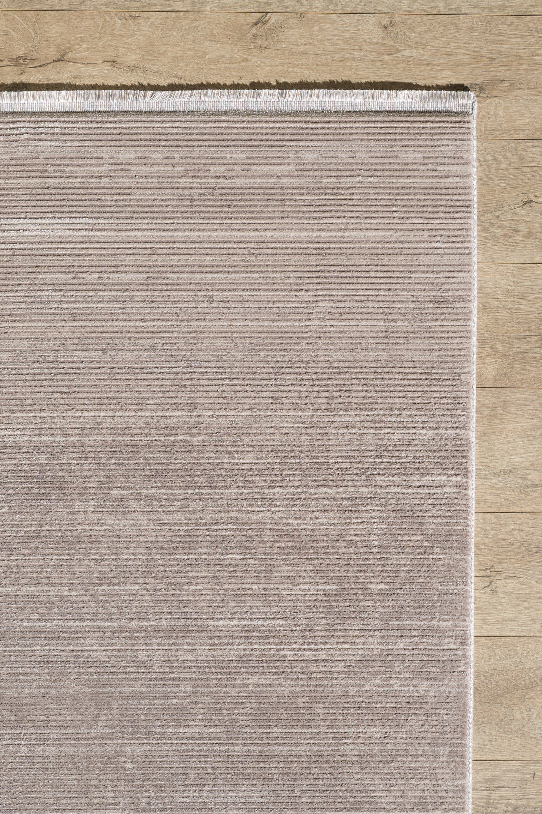 A close-up of the Earthy Textures Neutral Rug - M497J shows its rectangular, light brown minimalist design with subtle horizontal lines and a fringed top edge, placed on a light wooden floor.