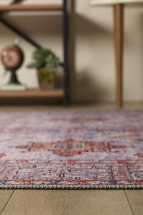 Bild 1 in Galerieansicht laden. A close-up of the Echelon Bordered Washable Rug - LCC3018, featuring vibrant red, purple, and orange patterns on a wooden floor; a plant, clock, and wooden furniture appear blurred in the background—ideal for high-traffic areas.