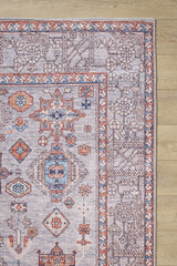 The Echelon Bordered Washable Rug - LCC3018, featuring geometric and floral patterns in blue, orange, and ivory, is perfect for high-traffic areas—only its corner is visible on a light wood floor.