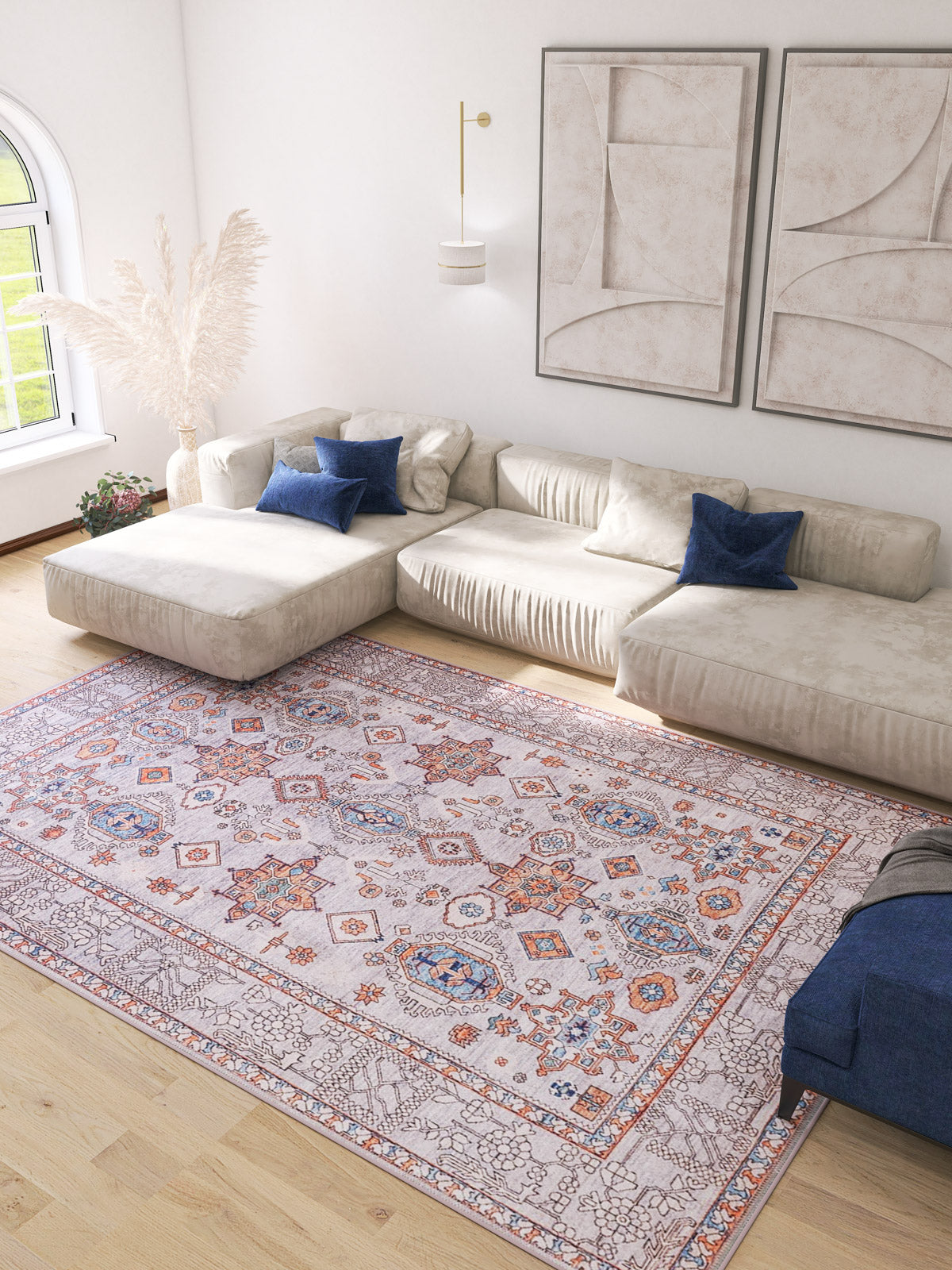 A bright living room with a large cream sectional, blue accent pillows, abstract beige wall art, light wood floors, and the Echelon Bordered Washable Rug - LCC3018 (Custom Size), perfect for high-traffic areas under sunlight from an arched window.