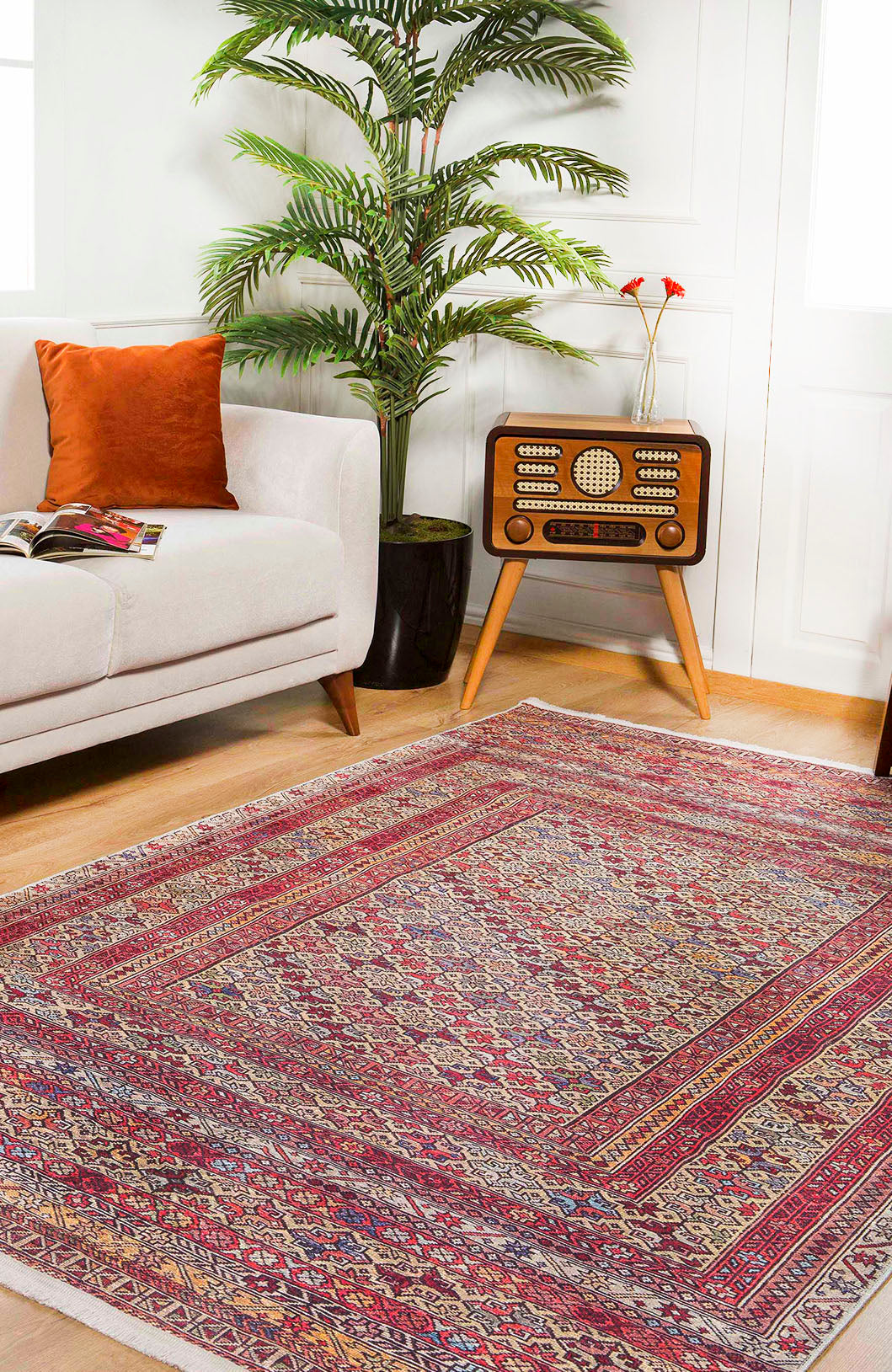 A cozy living room with the Elegant Embellishments - Washable Rug - JR43 (Custom Size) in red for high-traffic areas, a white sofa with an orange cushion, a vintage radio on wooden legs, a green potted plant, and a magazine on the sofa.