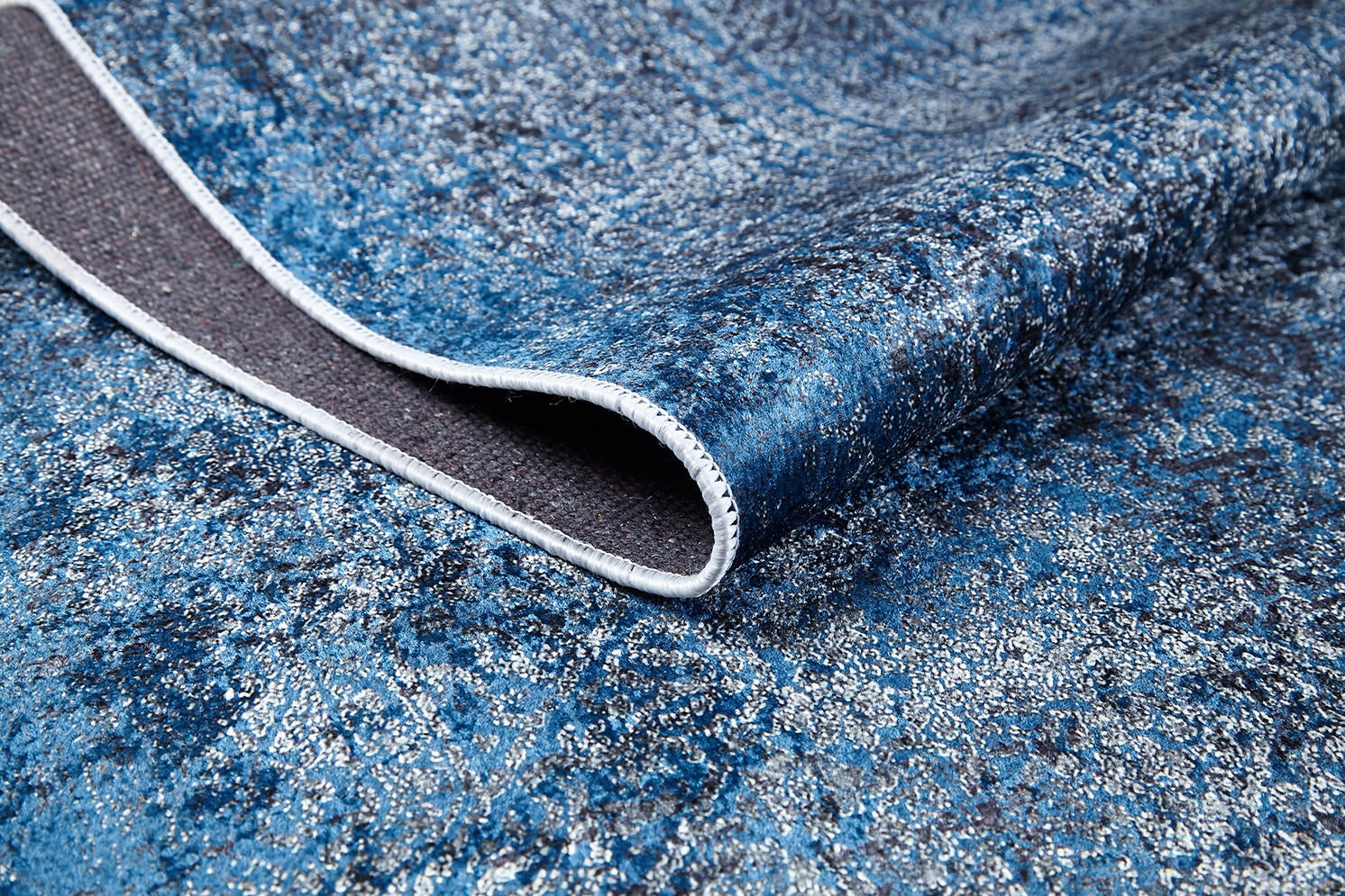 A close-up of the Elegant Vine - Washable Rug - JR1847 (Custom Size) shows its folded corner, detailed stitching, and durable backing. Light and dark blue tones with a subtle sheen make it ideal for high-traffic areas.