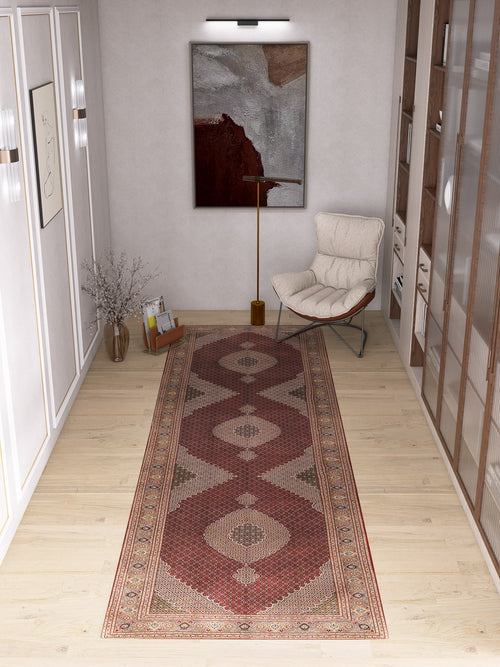 Load image 1 in gallery view. A modern hallway with light wood flooring showcases the Elysian Weave - Washable Rug - JRN1001 in red tones, a medallion design, an abstract wall painting, a cream lounge chair with a gold floor lamp, and a glass vase of dried branches.