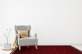 A cozy gray armchair with a beige pillow and blanket sits by a small stack of books and a plant, atop the Ember Stitch - Kilim - BHR02 red Turkish rug against a plain white wall in a minimally decorated living room.