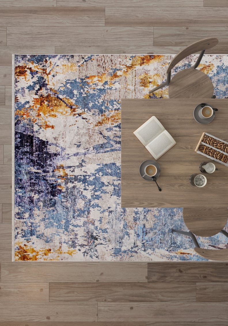 A top-down view of a modern dining table with two chairs sits on the Enigmatic Elegance - Washable Rug - JR787 (Custom Size); coffee, a book, and snacks rest on the table. The light wood floor adds warmth—perfect for high-traffic spaces.