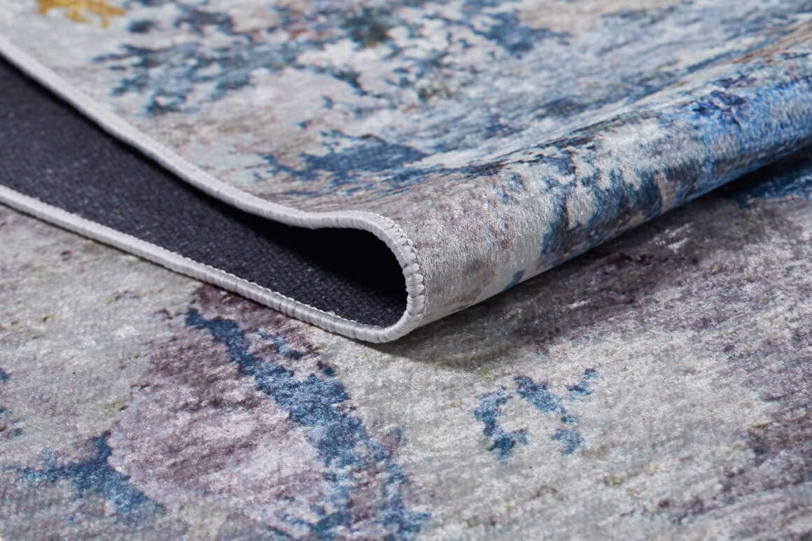 A close-up of the Enigmatic Elegance - Washable Rug - JR787 (Custom Size) shows its folded corner, revealing textured blue, gray, and beige tones—durable and washable for high-traffic areas, with a glimpse of its dark underside.