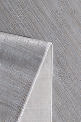 A close-up of the Essential Weave Modern Grey Rug - EW2836 (Outlet) US shows its minimalist design with subtle diagonal lines. One corner is flipped, revealing the underside and white edge trim against a matching patterned background.