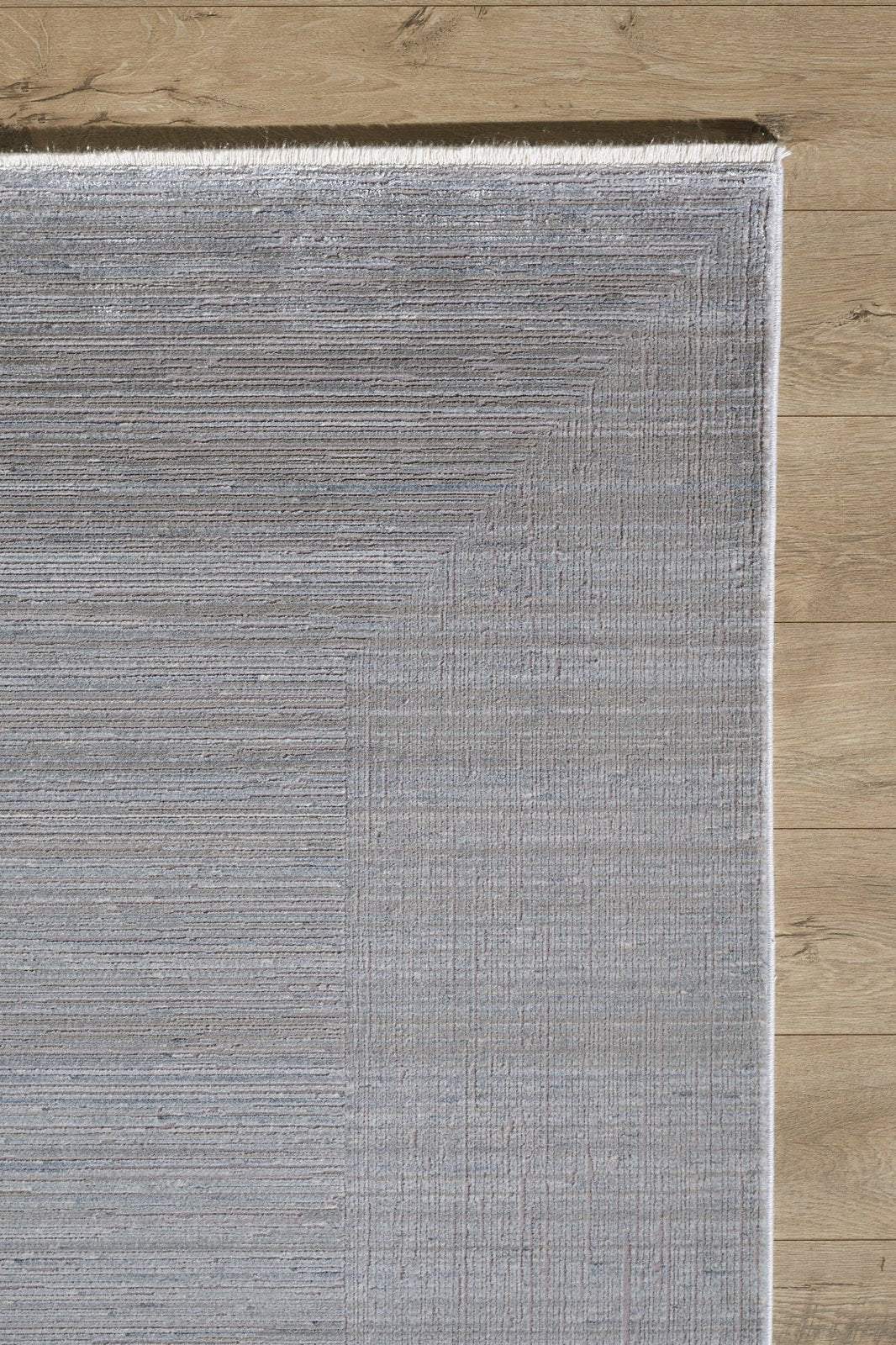 A close-up of the Essential Weave Modern Grey Rug - EW2836 (Outlet) US on a wooden floor, showing its light gray color, subtle linear design, and neatly finished edges.