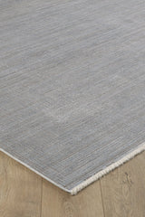 A close-up of the Essential Weave Modern Grey Rug - EW2836 (Outlet) US, a rectangular machine-made rug in a neutral grey with subtle textured patterns and short fringes, displayed on a natural-finish wooden floor with visible grain.