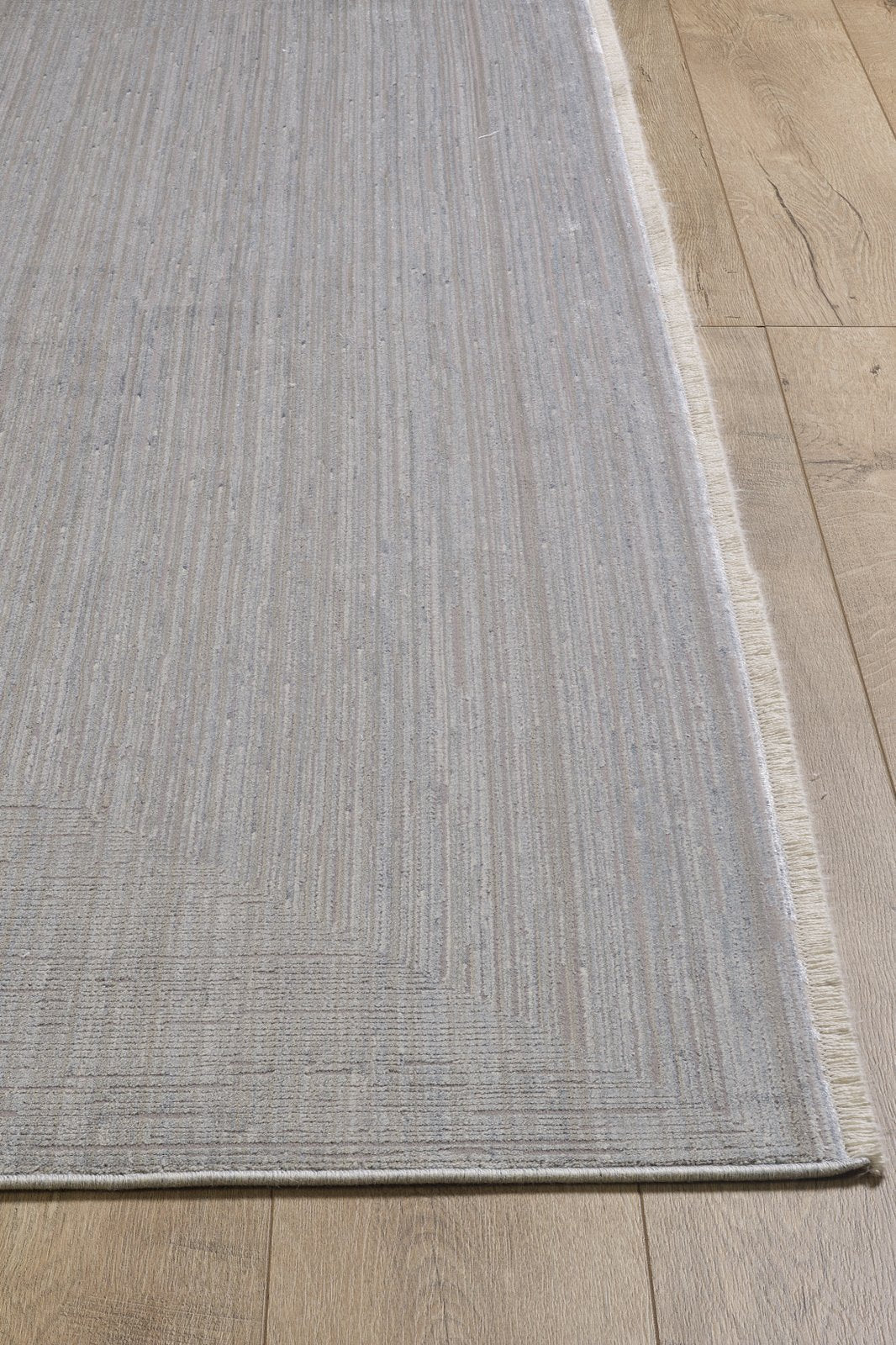 The Essential Weave Modern Grey Rug - EW2836 (Outlet) US is a large, rectangular, machine-made rug in a neutral grey with subtle stripes, soft texture, and slightly fringed edges, shown on a wooden floor for a minimalist look.