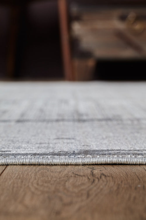 Load image 1 in gallery view. A close-up side view of the Essential Weave Modern Grey Rug - EW2836 (Outlet) US on a wooden floor, with a blurred background featuring part of a wooden furniture piece.