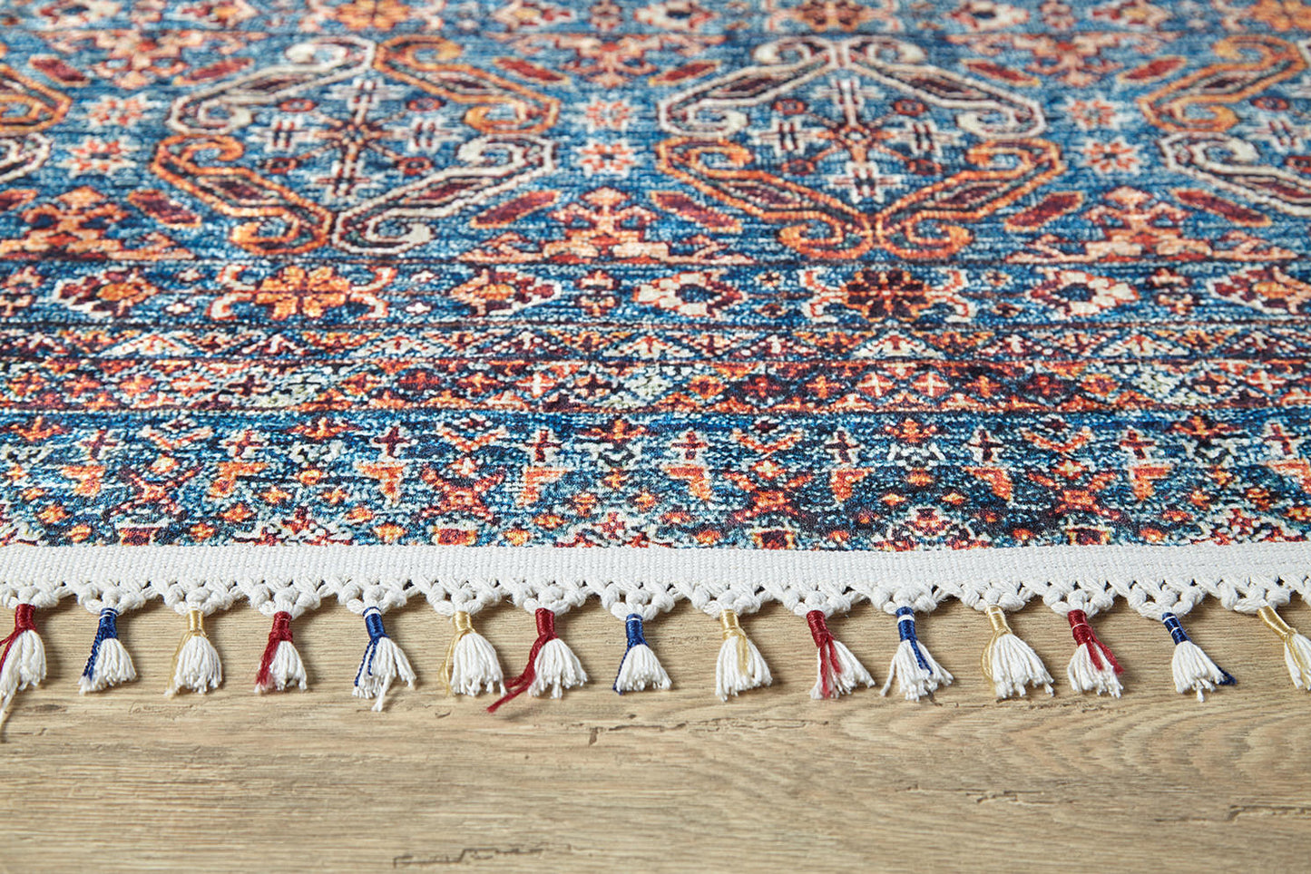 A close-up of the Ethereal Essence - Washable Rug - JR5087 (Custom Size), showcasing intricate blue, red, orange, and white geometric and floral designs with tassels—ideal for high-traffic areas on light wooden floors.