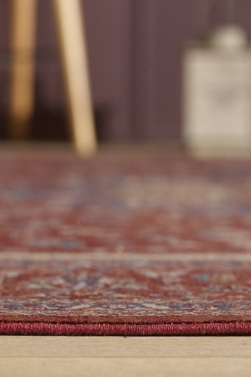 Load image 1 in gallery view. Close-up of the Ethereal Garden Medallion Rug - Burgundy - 2222A on a wooden floor, with its edge in focus and a blurred background featuring part of a wooden object and an indistinct rectangular item.