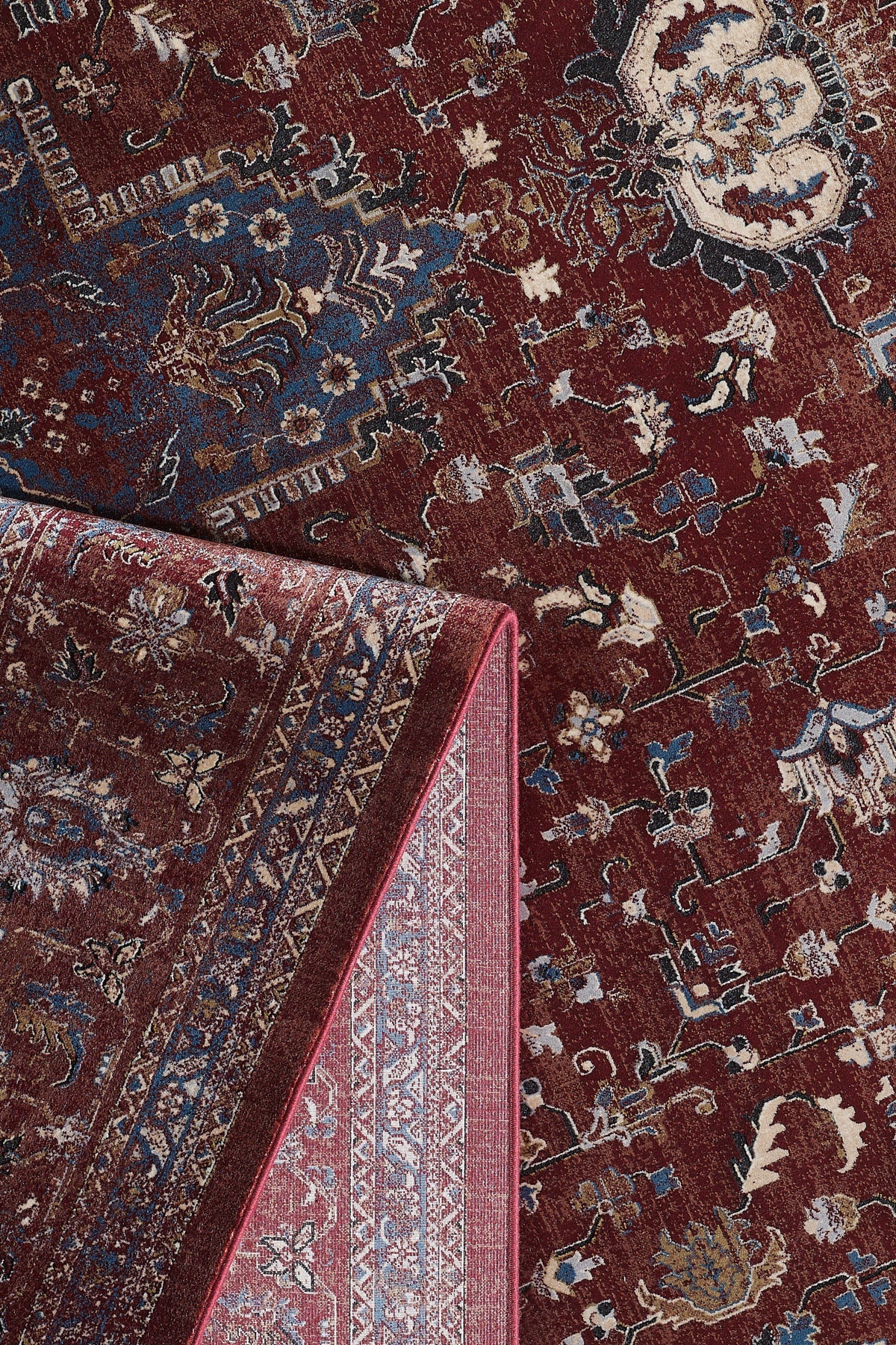 The Ethereal Garden Medallion Rug - Burgundy - 2222A features intricate cream and tan floral and geometric patterns on a richly patterned burgundy and blue background, with a corner folded to reveal its detailed underside and border.