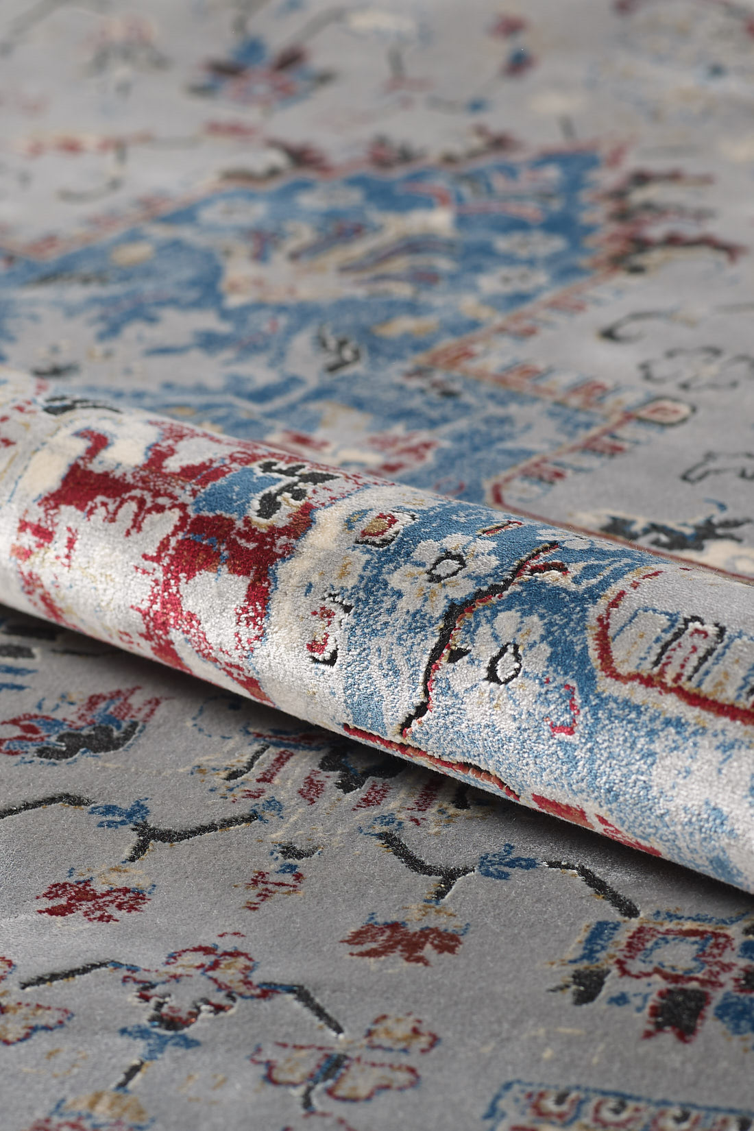 A close-up shows the rolled corner of the Ethereal Garden Medallion Rug - Grey - 2222C, featuring abstract red, blue, and beige designs on a soft gray background. The textured weave and detail of this grey and blue rug are clearly visible.