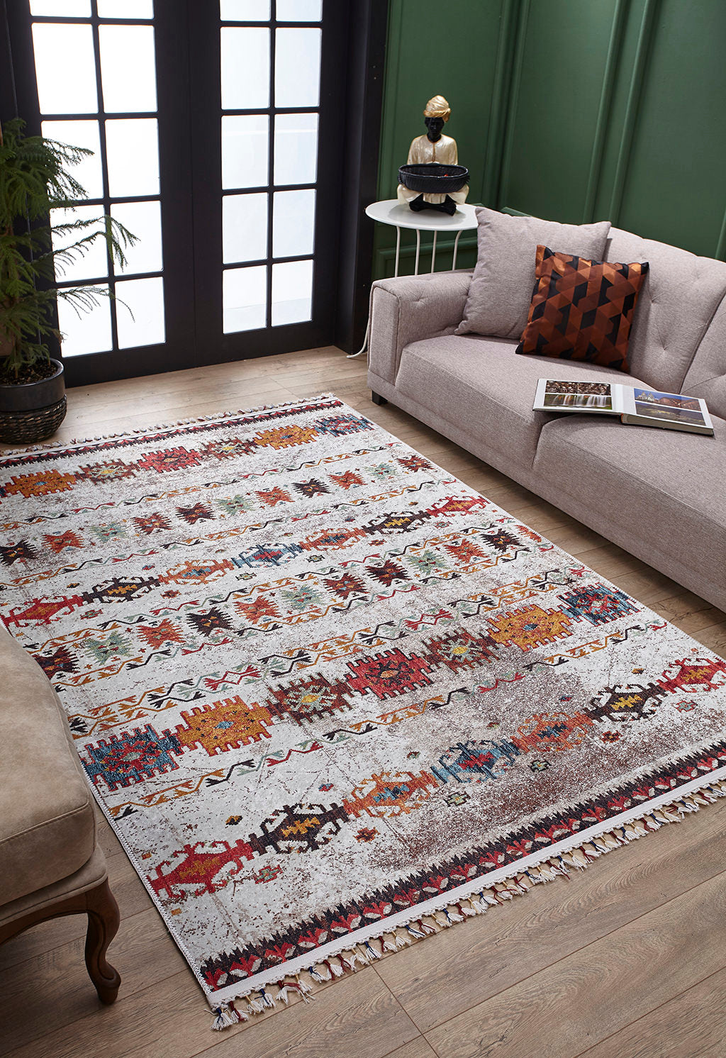 The Ethno Expanse - Machine-Washable Rug - JR5046 (Custom Size) adds color and texture to a living room with a gray sofa, brown chair, round side table, and green paneled walls on a wooden floor near tall windows.