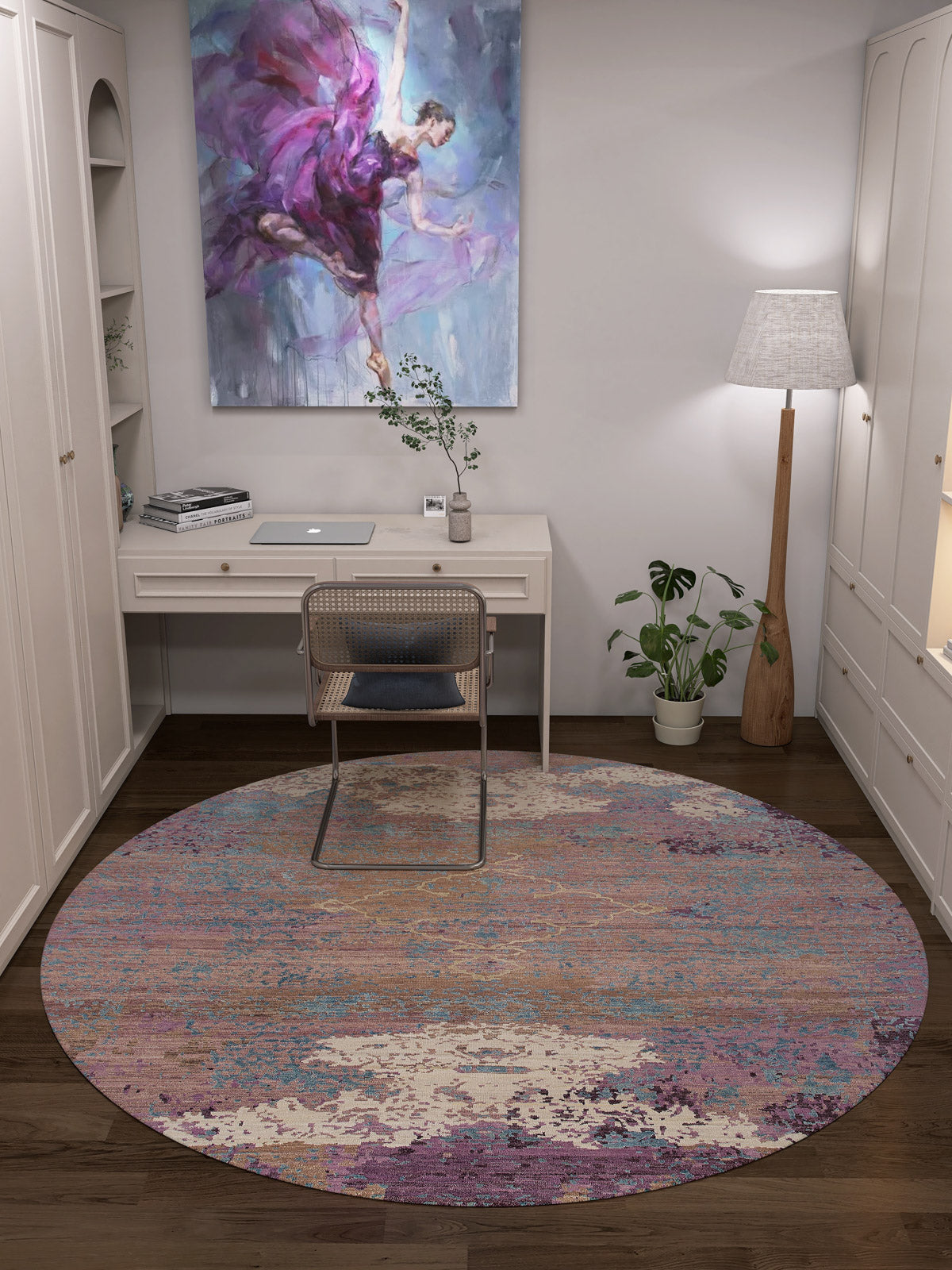 A home office features a white desk, laptop, rattan chair, Faerie Weave - Washable Rug - JRS005, and floor lamp. Art of a dancing woman hangs above the desk; a plant and books add charm. White cabinets and dark wood floors suit high-traffic areas.