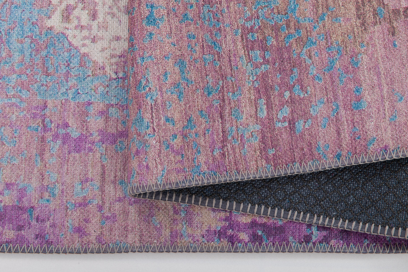 A close-up of the Faerie Weave - Washable Rug - JRS005, featuring a pink, purple, and blue abstract pattern with detailed edge stitching and a dark textured backing—ideal for high-traffic areas.