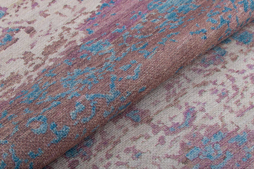 Load image 1 in gallery view. A close-up of the Faerie Weave - Washable Rug - JRS005 (Custom Size) highlights its purple abstract pattern with pink, blue, and beige tones, a folded section revealing woven detail—ideal as a washable rug for high-traffic areas.