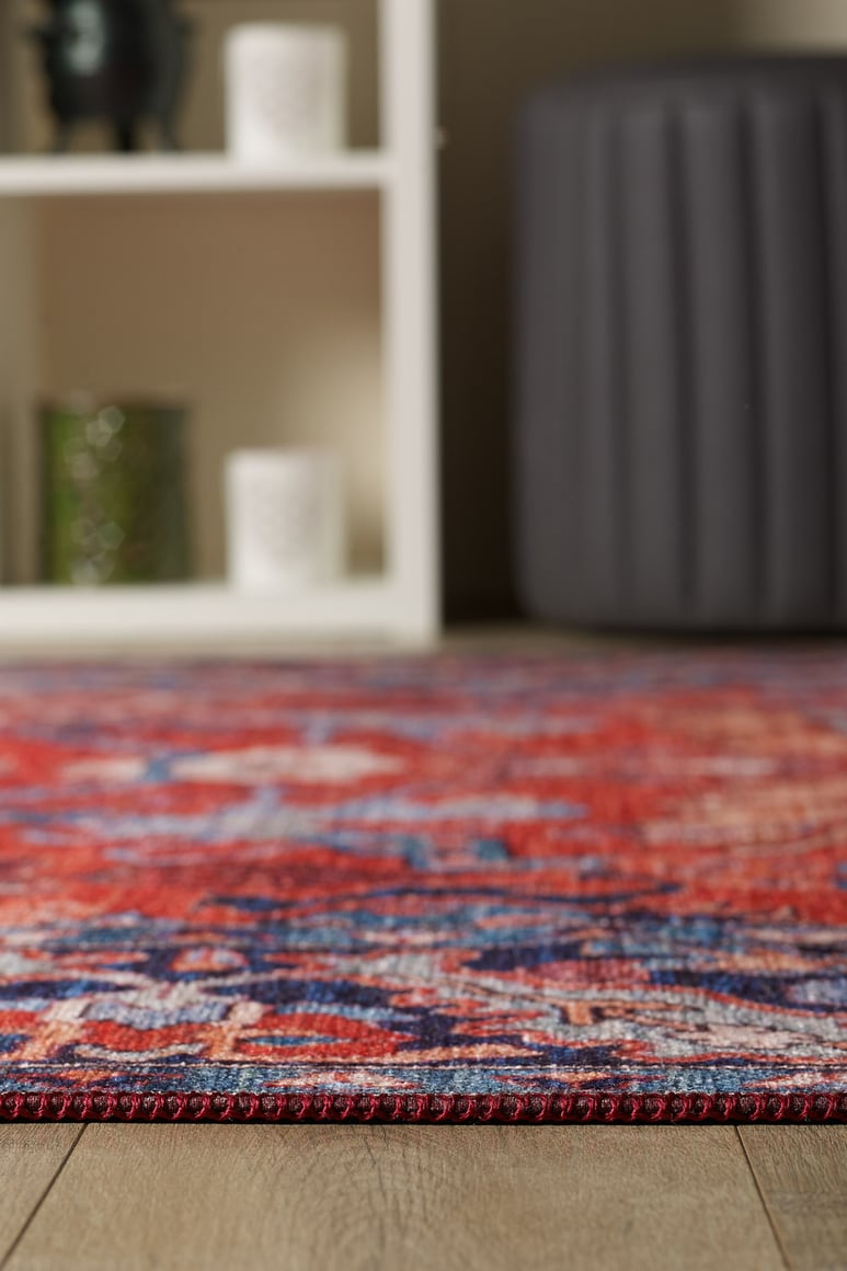 A close-up of the Galea Turkish-Design Coral Washable Rug (LCC3027, custom size) on a wooden floor, perfect for high-traffic areas, with a blurred white decor shelf and a dark gray round ottoman in the background.