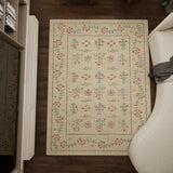 The Garden Cream - Washable Rug (JRN1026), a rectangular beige rug with red, green, and black floral patterns, is placed on a dark wooden floor beside a white curved sofa and a light-colored cabinet.