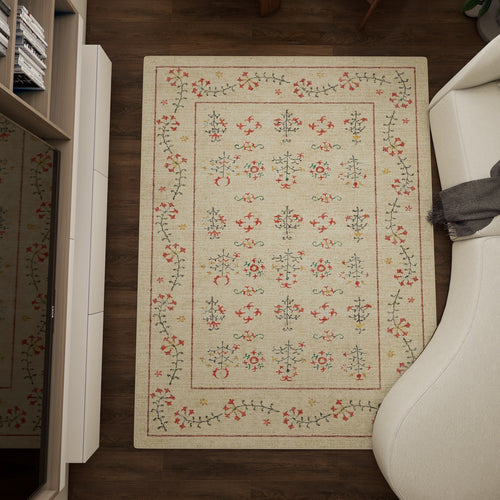 Load image 1 in gallery view. The Garden Cream - Washable Rug (JRN1026), a rectangular beige rug with red, green, and black floral patterns, is placed on a dark wooden floor beside a white curved sofa and a light-colored cabinet.