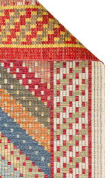 The Garnet Print - Kilim - NMD10 rug features a folded corner, showcasing its colorful low-pile polyester surface with red, yellow, blue, orange, and white geometric patterns in checkerboard and diagonal stripes.
