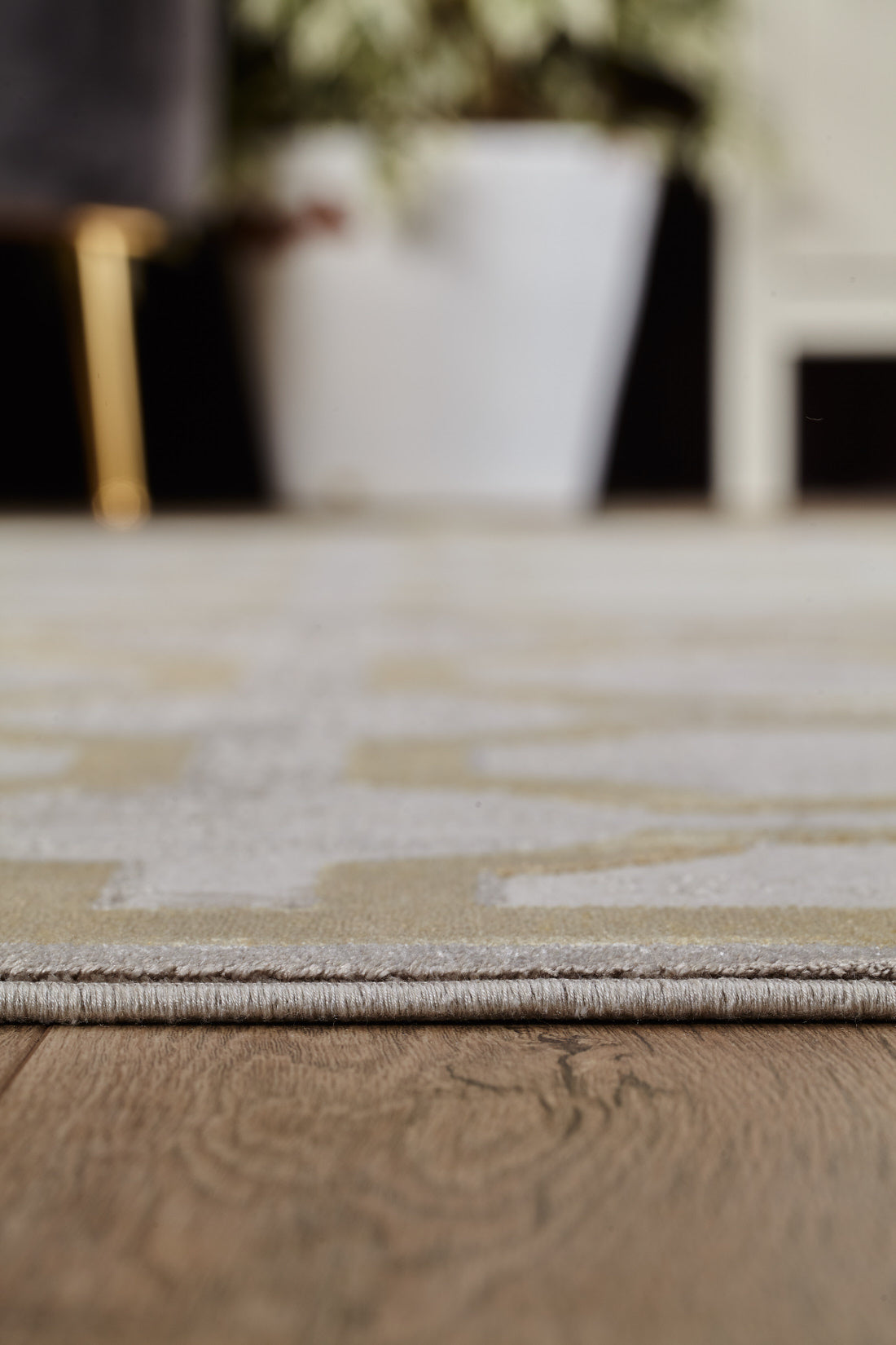 Close-up of the Geometric Down Mid Century Modern Rug - M534B, a contemporary beige and cream machine-made piece with a subtle lattice pattern on wooden flooring, near a large white plant pot against a blurred background.