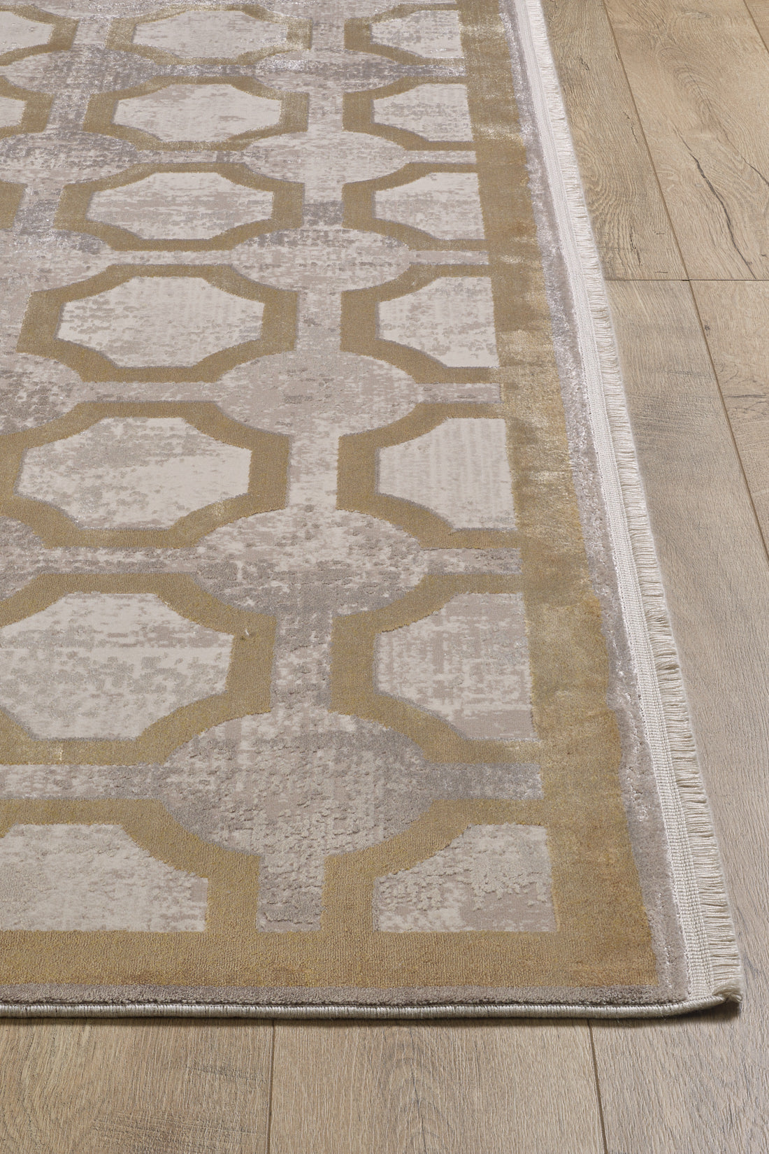 A close-up of the Geometric Down Mid Century Modern Rug - M534B shows its beige and gray lattice pattern, decorative border, and fringe on one edge, placed atop a light wood floor. This contemporary, machine-made rug adds modern style to any space.