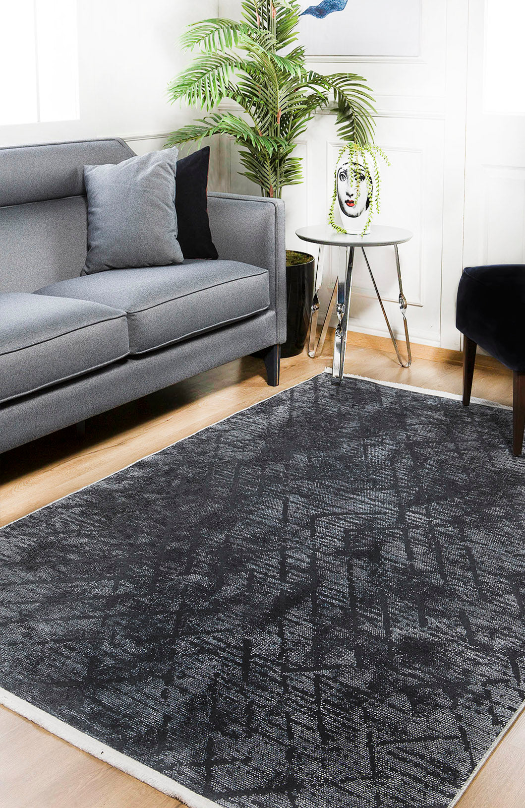 A modern living room with a gray sofa, black chair, Geometric Gems - Washable Rug - JR1342 (Custom Size) in black and gray—ideal for high-traffic areas—round white side table, decorative vase, green potted plant, and light wooden flooring.