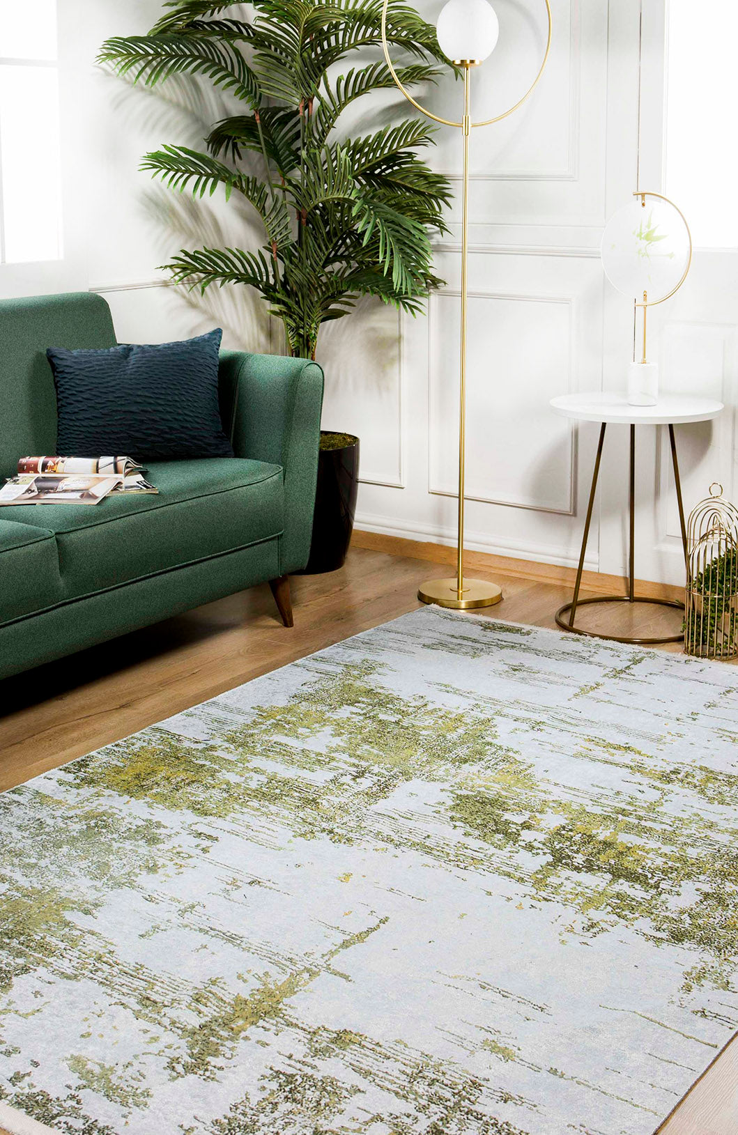 A modern living room with a green sofa, navy pillow, magazines, potted plant, marble side table and lamp, gold floor lamp, and the Geometric Geometry - Washable Rug - JR1290—perfect for high-traffic wooden floors with its abstract pattern.