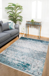 A modern living room has a gray sofa, decorative pillows, the Geometric Glimmers - Washable Rug - JR1423 (Custom Size) in blue for high-traffic areas, a green potted plant, and a small vintage desk with a trailing plant on white walls and wood floors.