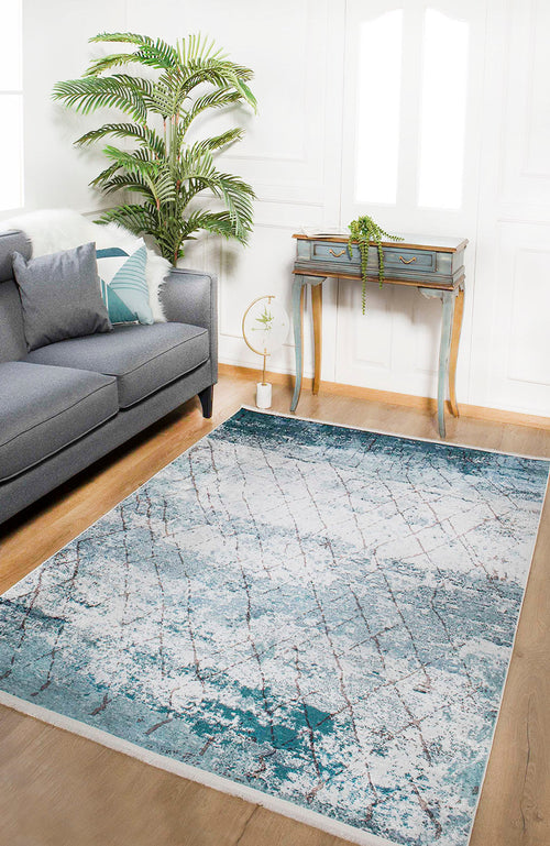Load image 1 in gallery view. A modern living room has a gray sofa, decorative pillows, the Geometric Glimmers - Washable Rug - JR1423 (Custom Size) in blue for high-traffic areas, a green potted plant, and a small vintage desk with a trailing plant on white walls and wood floors.