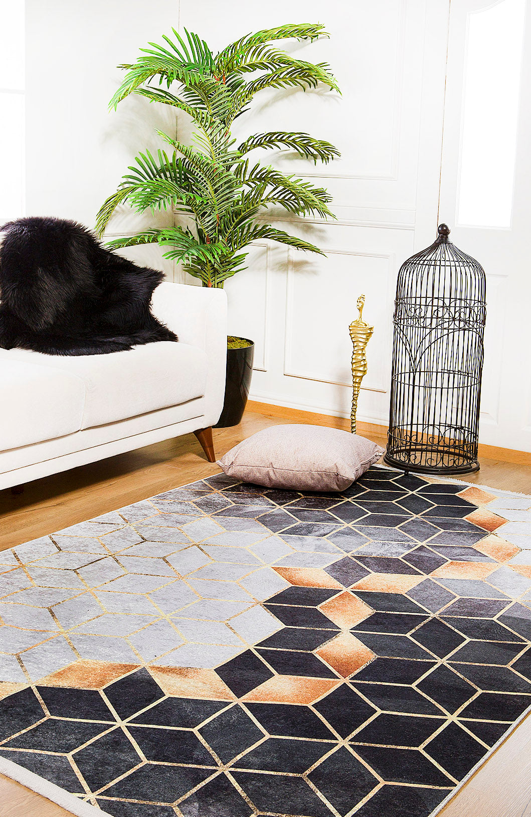 A modern living room featuring the Geometric Harmony - Washable Rug - JR1271 (Custom Size) in gray-anthracite, a white sofa with a black fur throw, a potted green plant, gold cobra decor, an empty black birdcage, and a beige floor cushion.