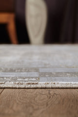 A close-up, low-angle shot of the Geometric Latte Premium Rug - M420G on a wooden floor, with blurred furniture and decor in the background, showcasing its minimalist design.