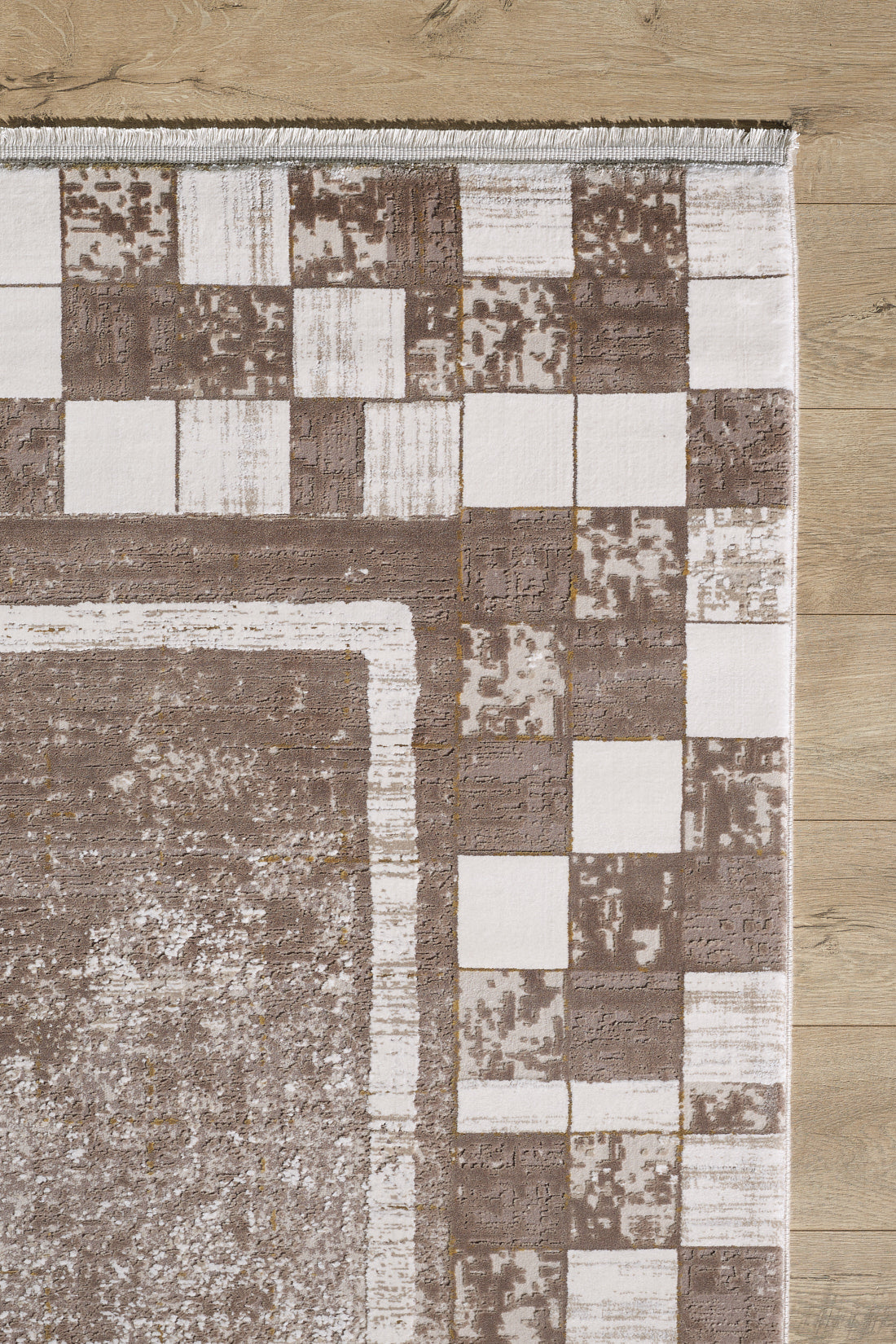 A close-up of the Geometric Latte Premium Rug - M420G shows its brown and white square border on a light wooden floor. This machine-made rug features a textured center, ideal for minimalist interiors.