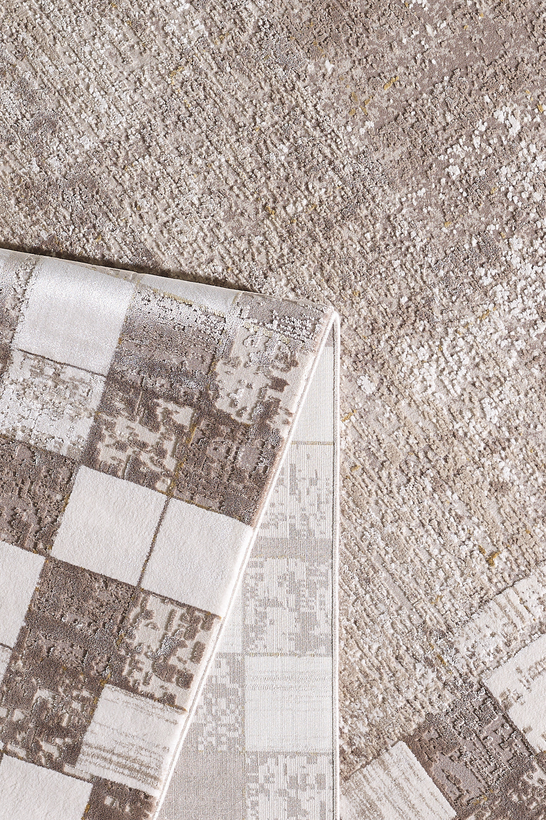 A close-up of the Geometric Latte Premium Rug - M420G shows a folded corner revealing its square geometric underside and textured beige and light brown surface, ideal for a modern, minimalist décor.
