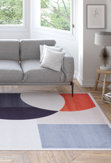 A modern living room with a light gray sofa, a dotted white pillow, the Geometric Zen Modern Washable Rug - MDJ002 (Custom Size) in navy, orange, and blue—perfect for high-traffic areas—a round table lamp, framed art, and large windows revealing green trees.