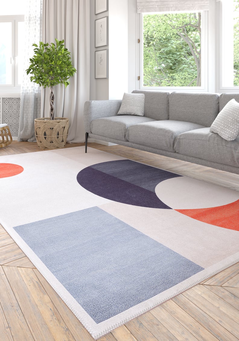 A modern living room with a gray sofa, white walls, and a large window features the Geometric Zen Modern Washable Rug - MDJ002 in blue, red, and purple tones—a stylish, durable choice for high-traffic areas.