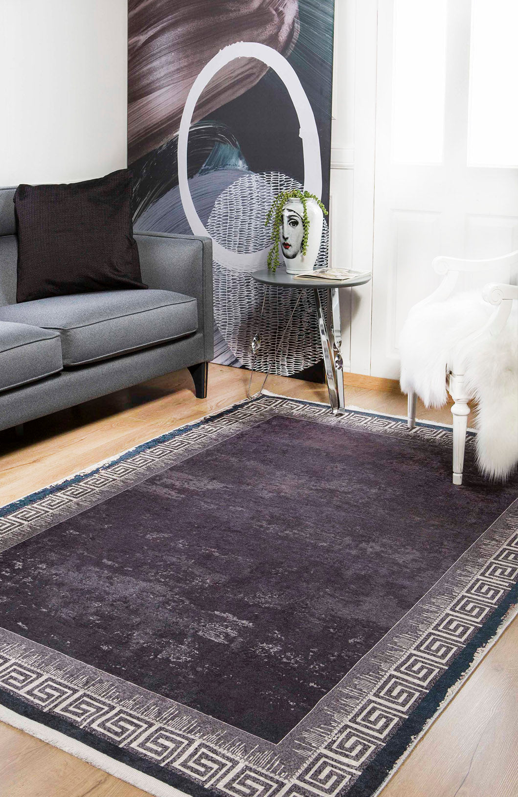 A modern living room with the Geometric and Chic - Washable Rug - JR1273 (Custom Size) in dark gray featuring a Greek key border, complemented by a gray sofa, black pillow, white chair with fur throw, side table, and decorative face vase.