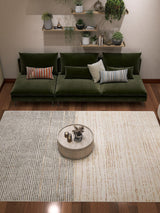 A modern living room features a green velvet sofa, mixed pillows, a round coffee table, and the Gilded Charm - Washable Rug - JRN26 (Custom Size), perfect for high-traffic areas. Wall shelves display potted plants and ornaments.