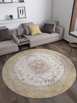 A modern living room features the Golden Oasis - Washable Rug - JR1930 in yellow, blue, and cream on dark wood floors. Nearby sits a light gray sectional sofa with assorted cushions and a wall shelf showcasing framed art and decor.