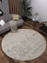 A modern living room features the Graceful Tapestry - Washable Rug - JR1975 on dark wood floors, a curved gray sofa with cushions and a throw, a wooden chair, a side table with a plant, and a tall houseplant in the corner.